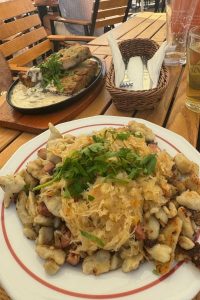 A meal from the Pyra Bar in Gdansk featuring a plate of fried dumplings with sauerkraut and herbs. The table also has a glass of beer and a basket with cutlery and napkins.