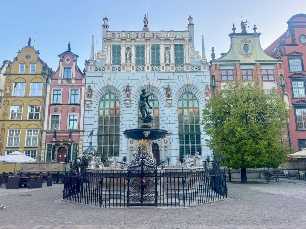 Neptune’s Fountain stands in Gdańsk’s Long Market, with the ornate white facade of Artus Court behind it, framed by historic townhouses and trees