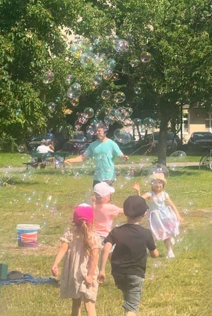 Children chasing giant soap bubbles beside the Vistula River in Krakow, with the historic Wawel Castle behind them.