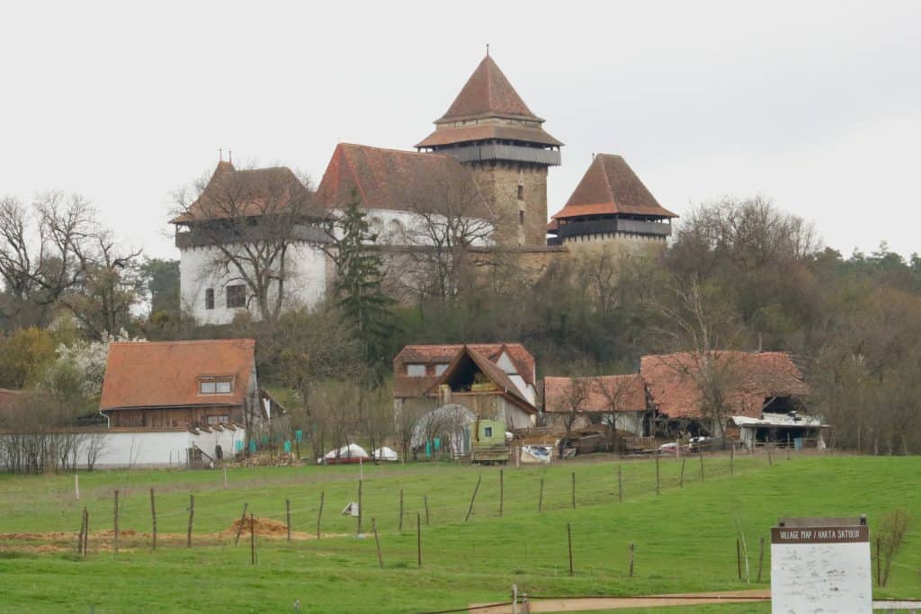 Fortified church in Viscri, Romania, with red-roofed towers and surrounding village homes, set on a grassy hill amid trees and rustic fencing.