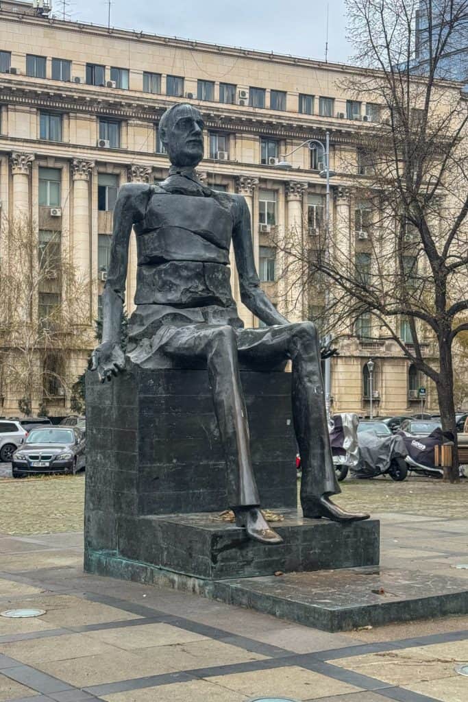 Modern bronze statue of a seated figure on a pedestal in Bucharest, set against an urban backdrop of historic architecture and parked cars.