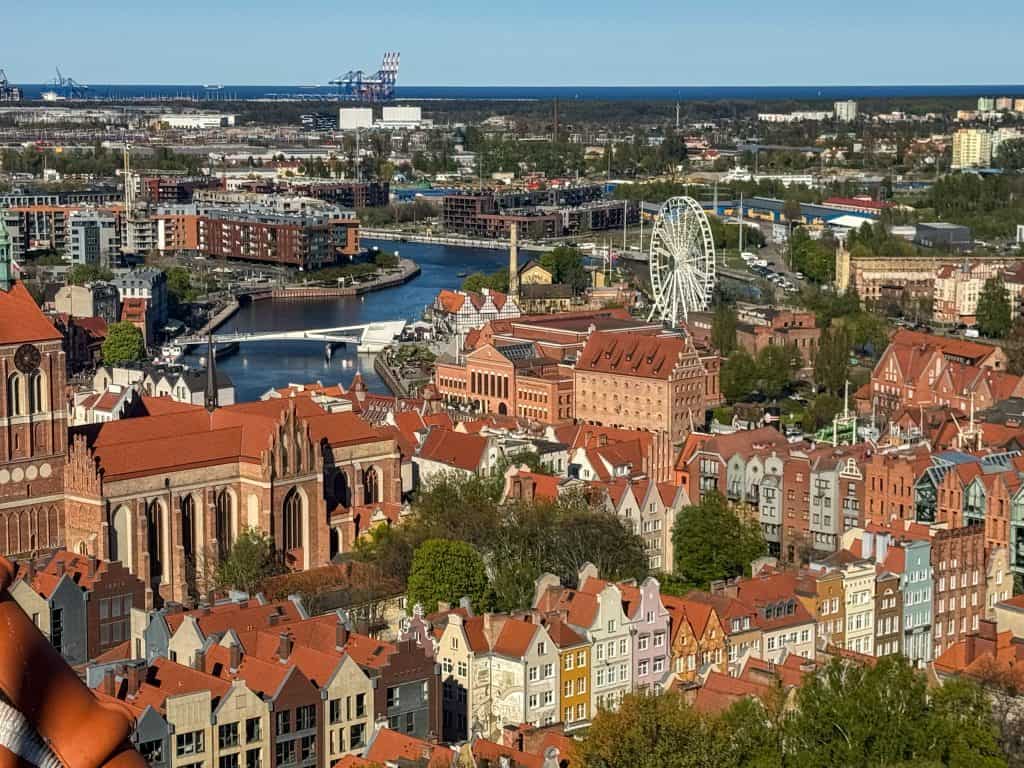 “Panoramic view over Gdańsk from St. Mary’s Tower, showing red-roofed Old Town, Motława River, ferris wheel, and green parks under a hazy sky.