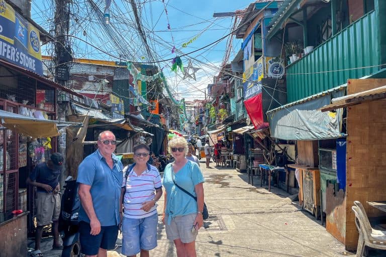 Three people standing in a lively Manila street lined with shops and colorful signs and tangled power lines overhead.