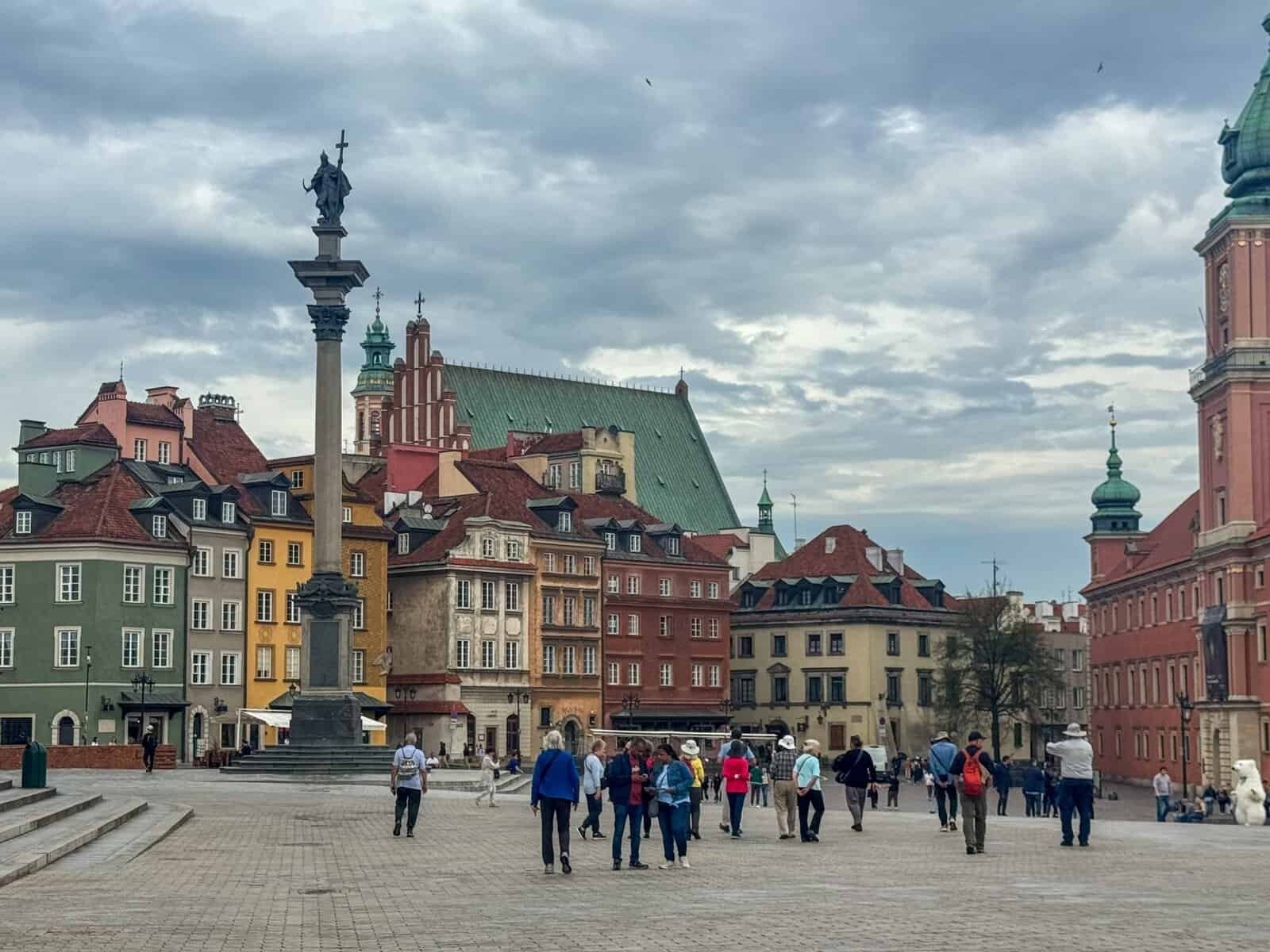 Sigismund's Column in Castle Square, Warsaw, featuring the tall Corinthian column topped with a bronze statue of King Sigismund III Vasa holding a cross and sword, surrounded by colorful historic buildings and pedestrians under a cloudy sky.