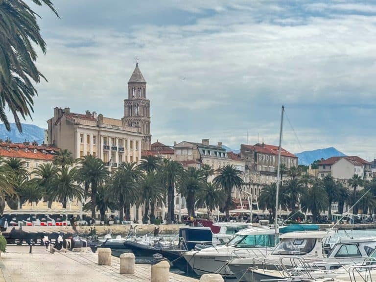Waterfront promenade in Split, Croatia, with palm trees, docked boats, and the bell tower of the Cathedral of Saint Domnius rising above historic stone buildings