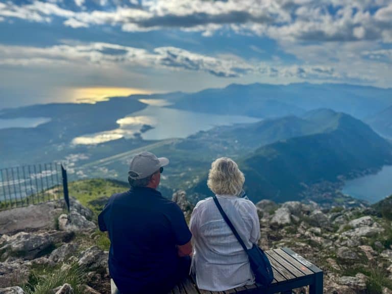 Two people on a bench overlooking Montenegro’s mountainous coastline and shimmering bay, under a partly cloudy sky—capturing the region’s peaceful, panoramic natural beauty.