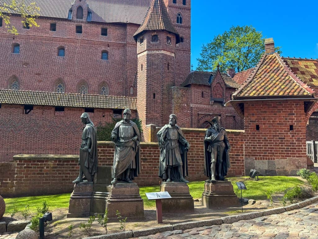 Four statues of historical figures stand in a row on pedestals in front of a red brick wall at Malbork Castle, Poland. The castle’s Gothic architecture—arched windows, tiled roofs, and fortress-like walls—are visible in the background.