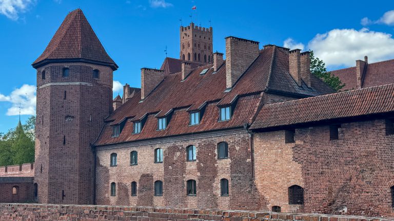 Exterior view of Malbork Castle’s red-brick tower and steep tiled roof under a blue sky, showcasing medieval architecture and fortress design.