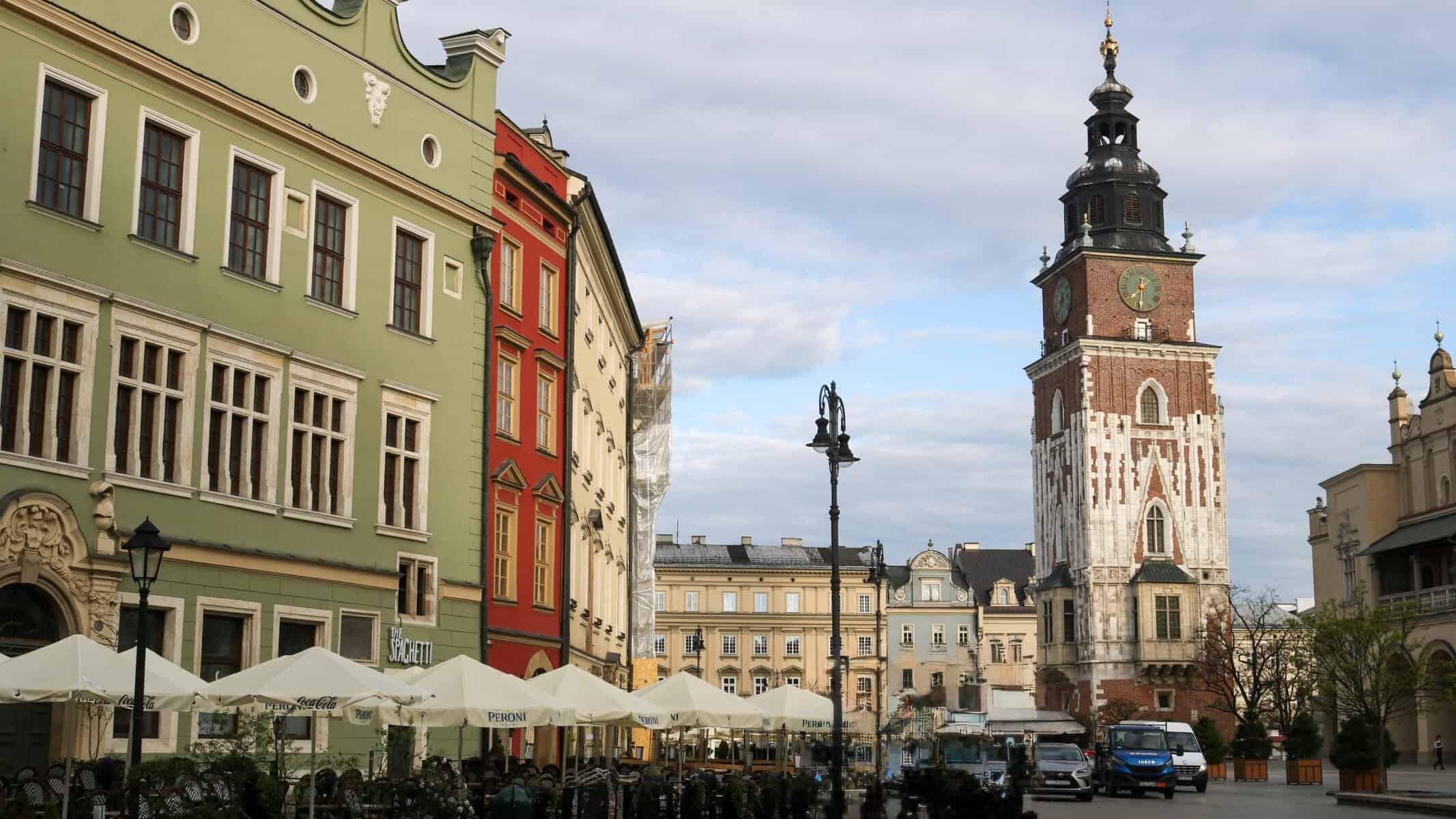 Historic street scene in Kraków’s Old Town, featuring colorful townhouses with ornate facades, outdoor café seating under white umbrellas, and the iconic spire of St. Mary's Basilica rising in the background beneath a partly cloudy sky