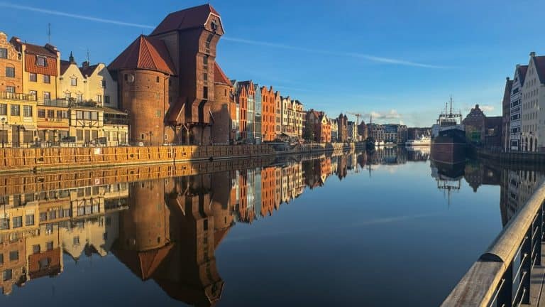 View of Gdańsk’s historic waterfront along the Motława River, featuring colorful gabled buildings, the medieval crane Żuraw, and a docked ship under a clear blue sky.