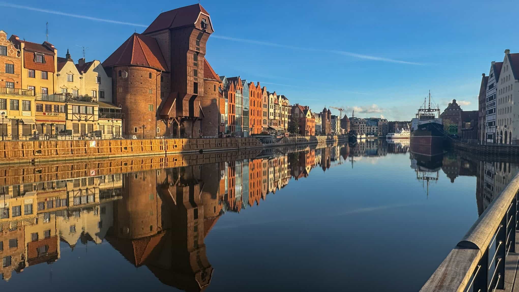 View of Gdańsk’s historic waterfront along the Motława River, featuring colorful gabled buildings, the medieval crane Żuraw, and a docked ship under a clear blue sky.