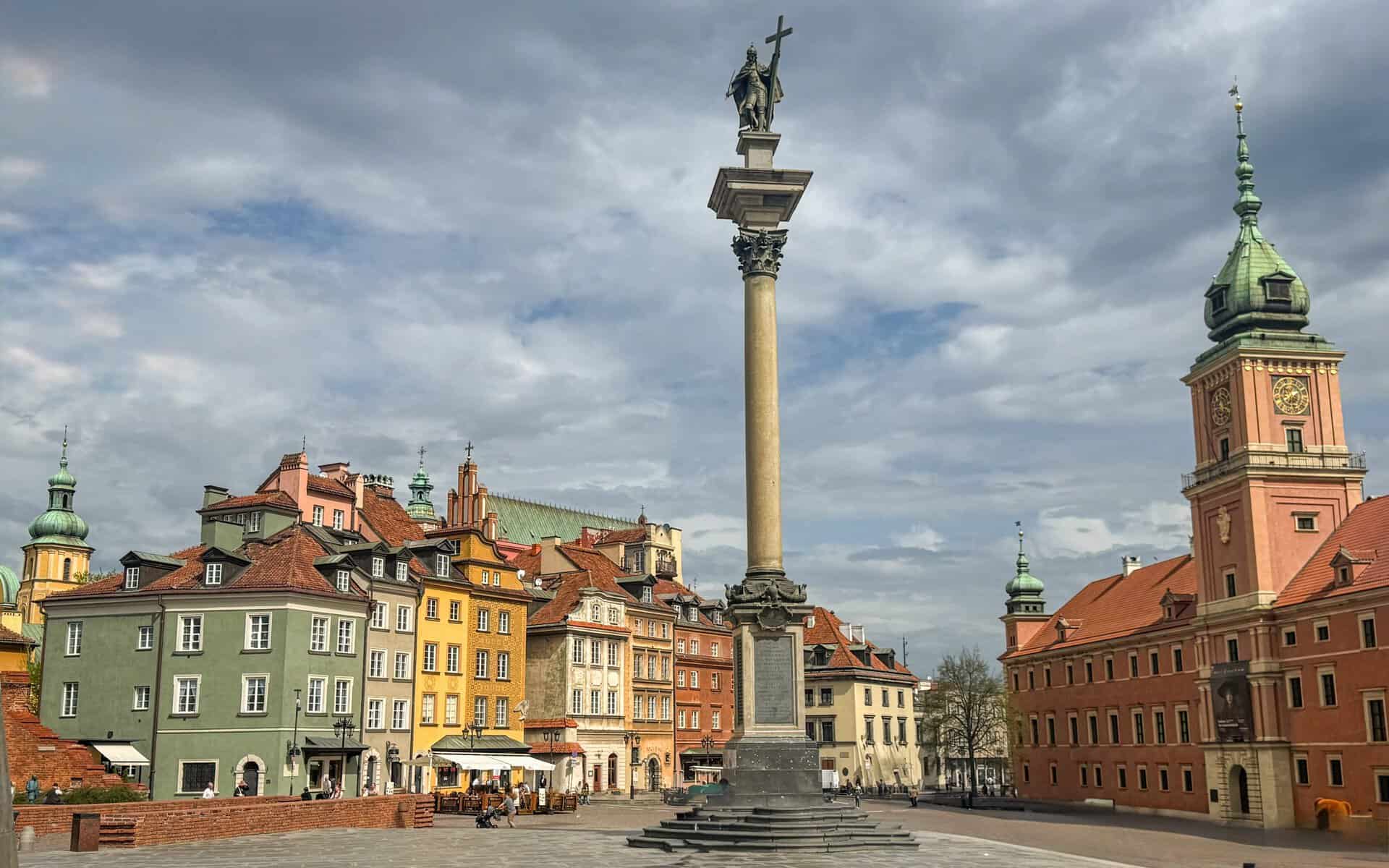 Sigismund’s Column in Warsaw’s Castle Square, featuring King Sigismund III Vasa holding a sword and cross atop a tall pedestal, surrounded by pastel-hued historic buildings under a partly cloudy sky.