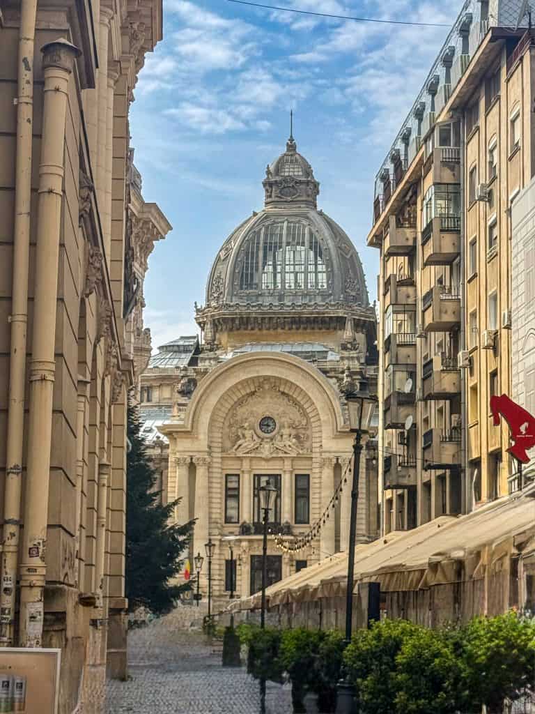 A grand Beaux-Arts bank building with a domed glass roof and ornate stone façade in central Bucharest. The narrow street is lined with classical buildings, outdoor cafés with white umbrellas, and planters filled with greenery under a partly cloudy sky.