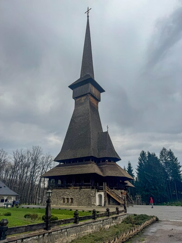 A tall wooden church with a steeply pitched roof and pointed steeple stands among trees under a cloudy sky, showcasing traditional Eastern European craftsmanship.