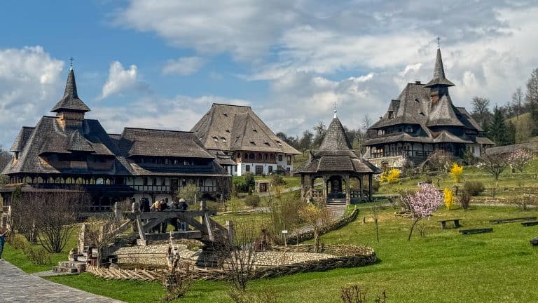 A serene monastery complex in Maramureș, Romania, featuring steeply pitched wooden roofs, carved timber structures, and blooming gardens. Stone paths wind through the lush greenery, connecting churches, gazebos, and traditional buildings