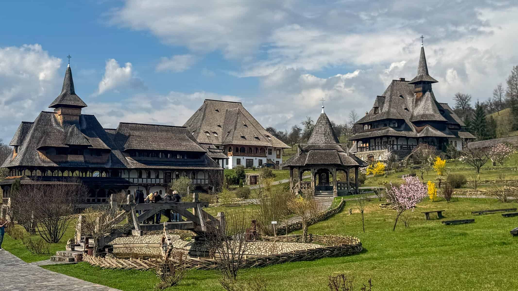 A serene monastery complex in Maramureș, Romania, featuring steeply pitched wooden roofs, carved timber structures, and blooming gardens. Stone paths wind through the lush greenery, connecting churches, gazebos, and traditional buildings