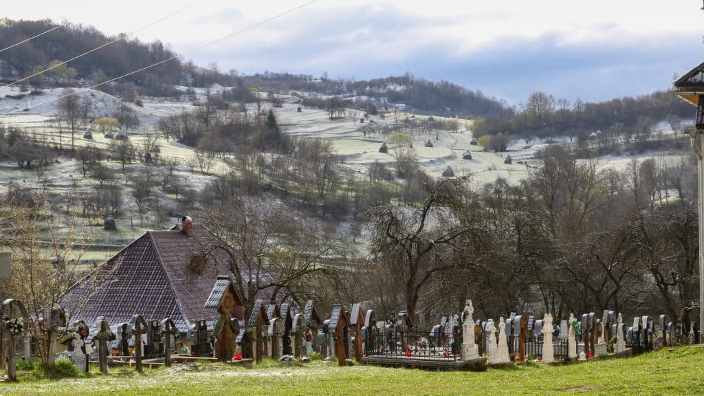 Snow-dusted cemetery in rural Maramureș, Romania, with traditional gravestones set against rolling hills and scattered trees under a partly cloudy sky.