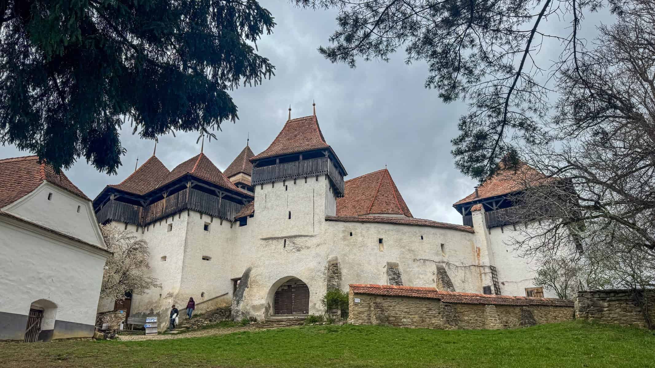 A fortified medieval church complex with red-tiled roofs and multiple towers, surrounded by stone walls. The structure sits on a grassy rise, framed by trees under an overcast sky.