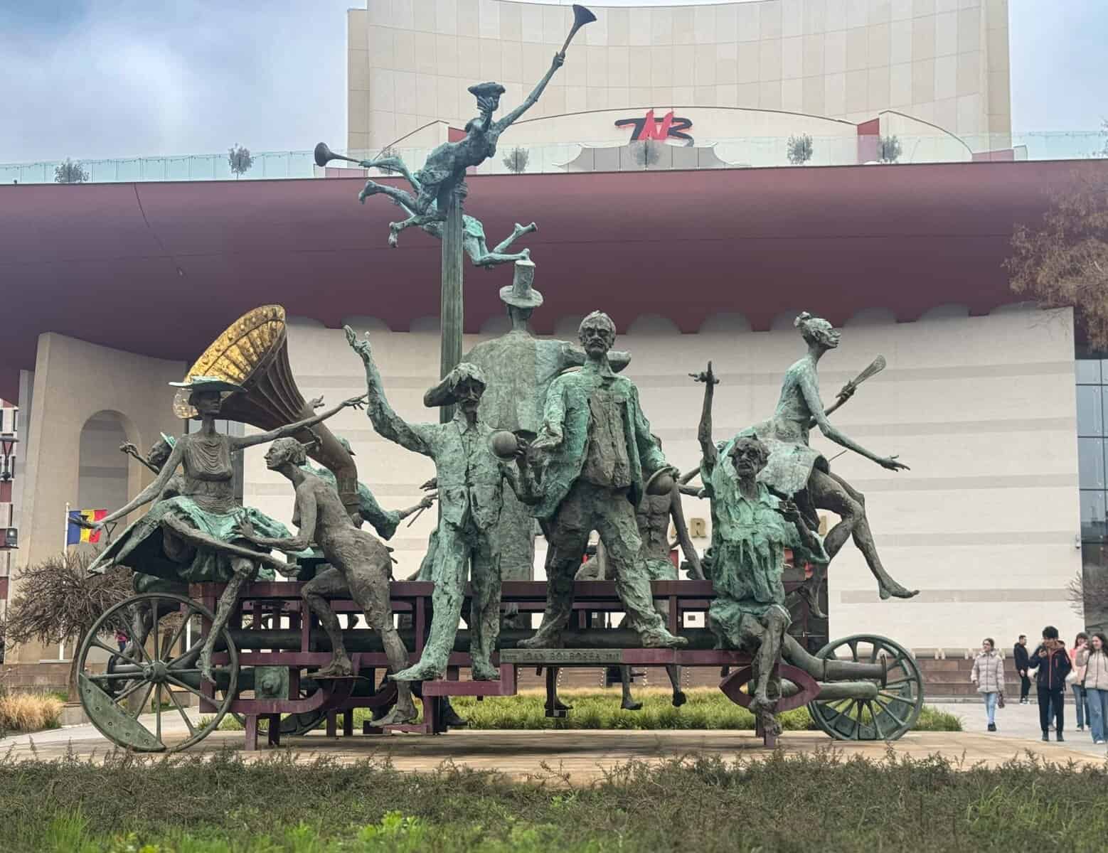 Bronze sculpture in front of the National Theatre Bucharest (TNB), featuring a lively group of human figures on a wheeled platform. The characters are posed dynamically—some playing instruments, others gesturing theatrically, with one figure performing a handstand on a pole. A large gramophone-like structure and architectural elements frame the scene.