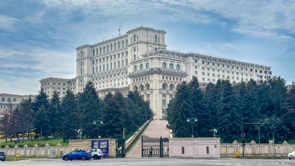 iew of the Palace of the Parliament in Bucharest, Romania, showcasing its massive neoclassical façade with symmetrical windows and ornate detailing. A gated entrance and parked cars appear in the foreground, framed by trees under a partly cloudy sky.