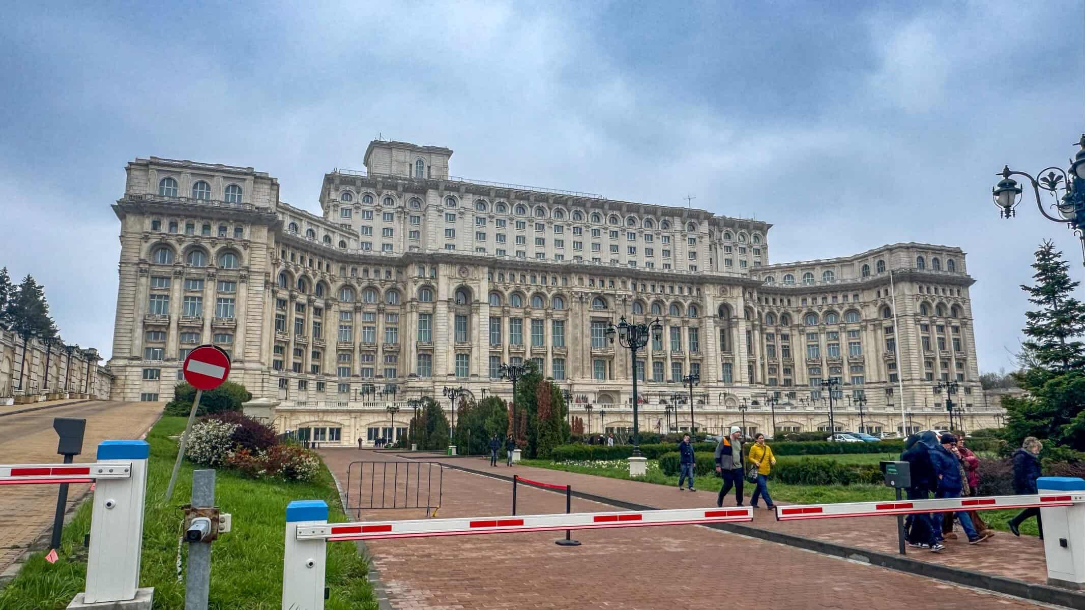 Front view of the Palace of the Parliament in Bucharest, Romania, showcasing its massive neoclassical façade with symmetrical rows of columns, arched windows, and ornate stonework. A red-and-white barrier gate and 'Do Not Enter' sign mark the paved foreground, bordered by trimmed bushes and flowerbeds.