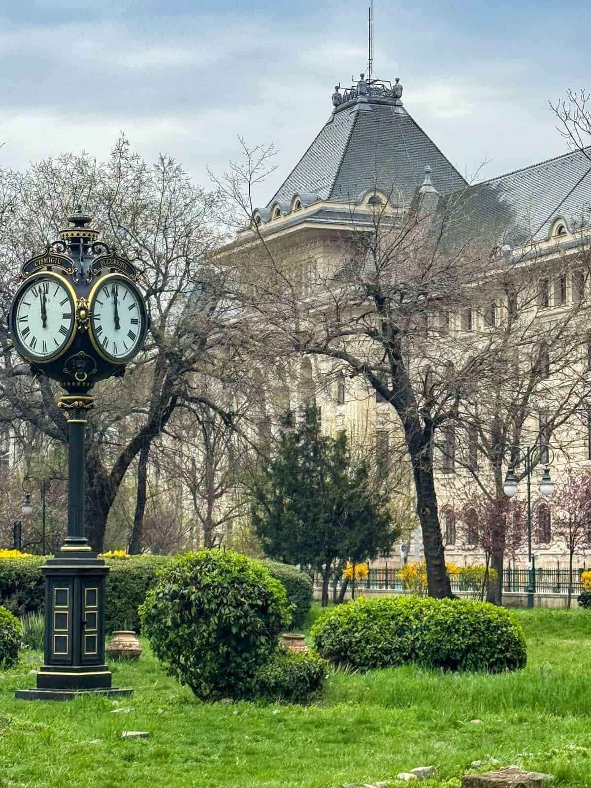 Urban garden in Bucharest featuring a vintage-style black and gold street clock with dual faces showing 11:50. The clock is surrounded by neatly trimmed bushes and trees, some bare, suggesting early spring or late autumn. In the background stands an ornate building with a steep roof and classical architectural details, adding historical charm to the peaceful park setting.