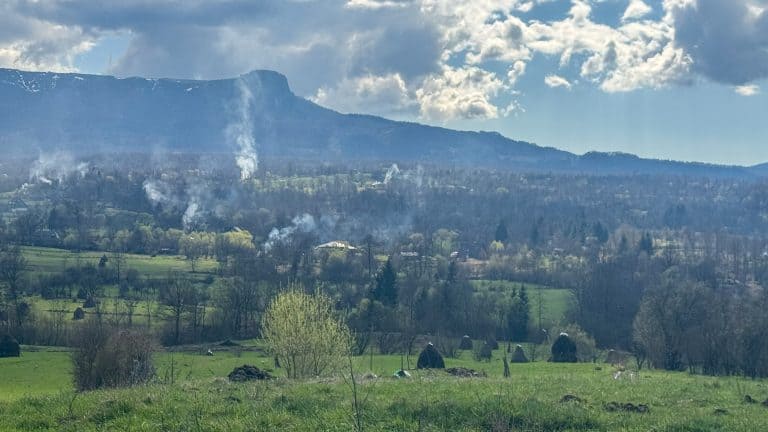 Smoke rising from scattered homesteads in a green valley leading into Breb, Romania, framed by a flat-topped mountain and early spring fields.