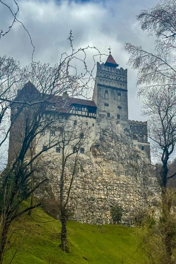 Bran Castle perched on a grassy hill in Romania, framed by leafless trees and a moody sky.