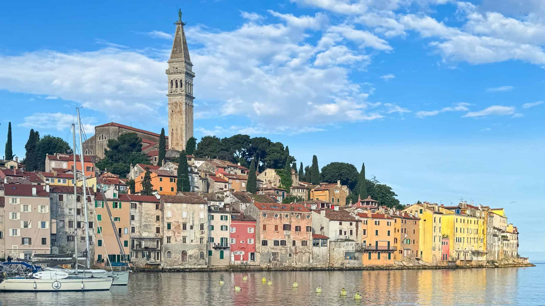 Waterfront view of Rovinj’s old town with pastel-colored buildings and a tall church bell tower rising above the harbor, sailboats moored in the calm blue water, and cypress trees on the hillside under a lightly clouded sky.