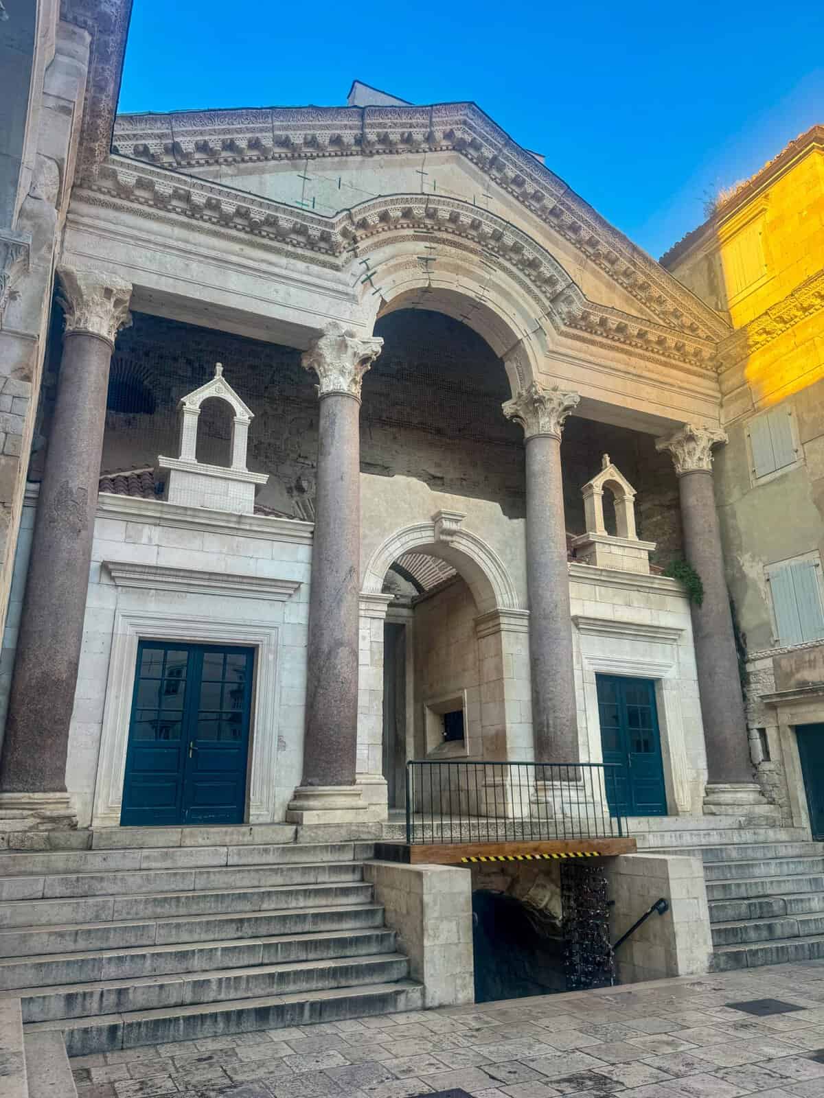 Stone façade within Diocletian’s Palace in Split, featuring ornate columns, arches, and statues, with two large doorways at the base under a clear blue sky.