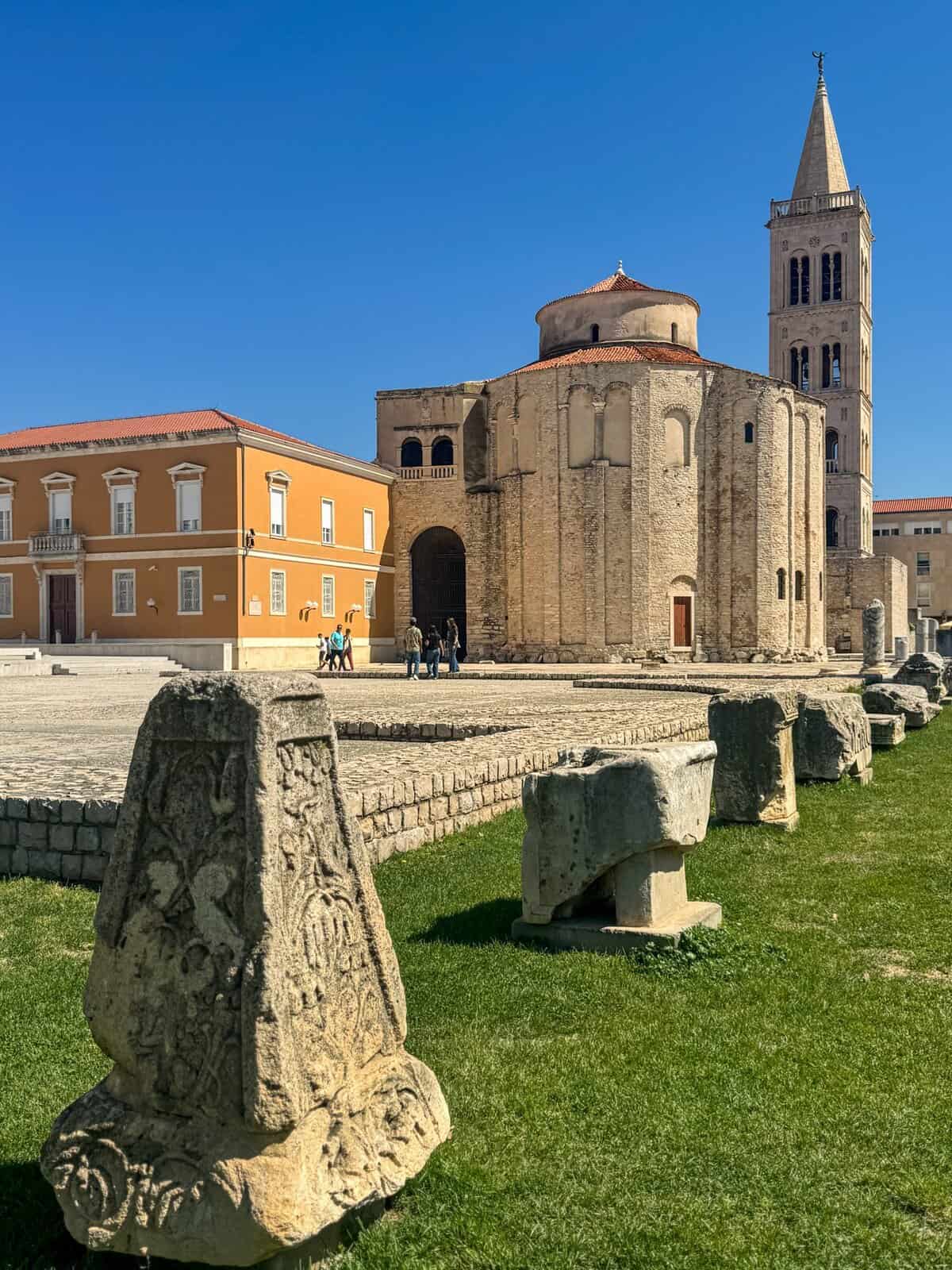 The round Church of St. Donatus and its tall Romanesque bell tower in Zadar, with ancient stone ruins displayed on a grassy area in the foreground under a clear blue sky.