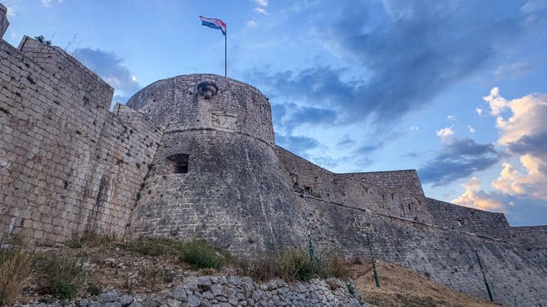 Fortica (Španjola) Fortress rises above Hvar Town, its stone walls and Croatian flag glowing in warm sunset light as it sits on a rocky hillside beneath a partly cloudy sky.