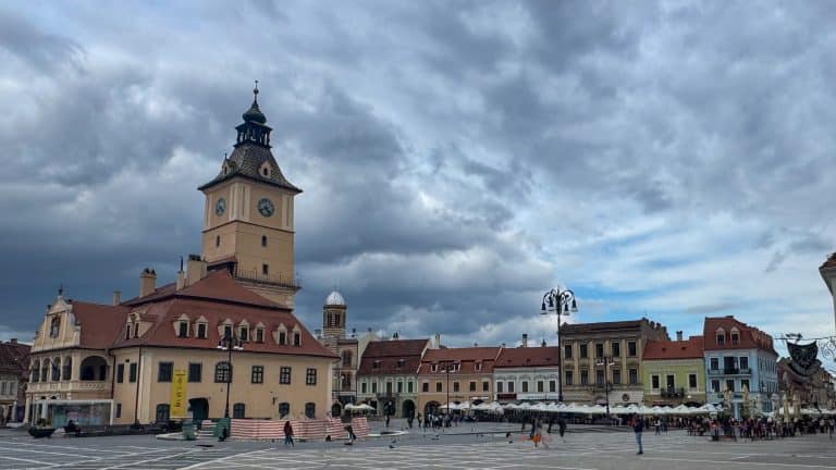 A wide view of Brașov’s Council Square with the historic Council House and its tall clock tower in the centre, surrounded by colourful medieval buildings with red‑tiled roofs. People walk across the patterned stone plaza under a dramatic, cloudy sky, with pigeons scattered around the square.