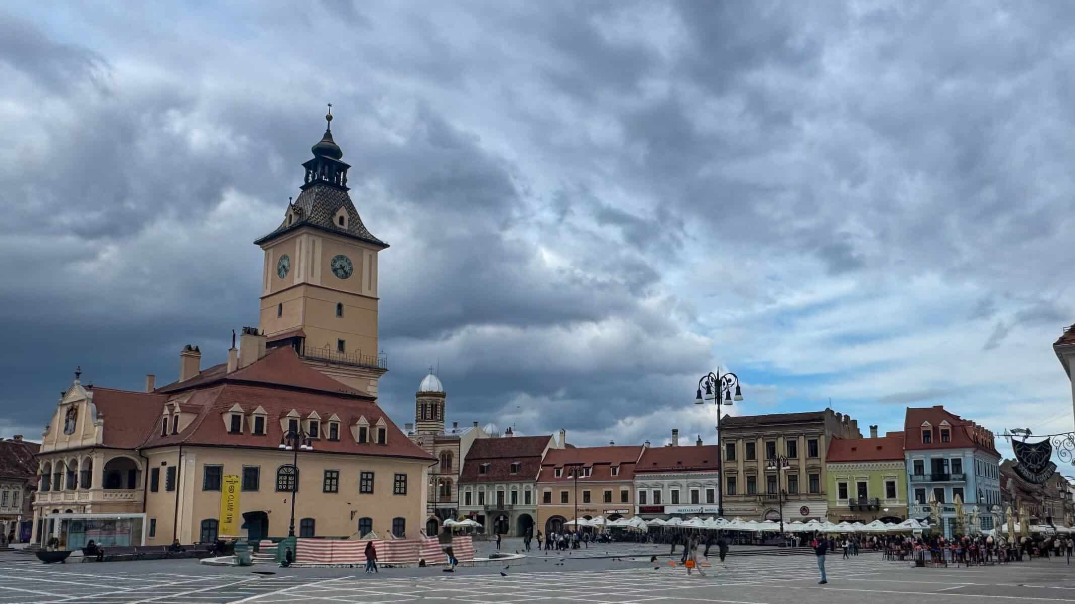 A wide view of Brașov’s Council Square with the historic Council House and its tall clock tower in the centre, surrounded by colourful medieval buildings with red‑tiled roofs. People walk across the patterned stone plaza under a dramatic, cloudy sky, with pigeons scattered around the square.
