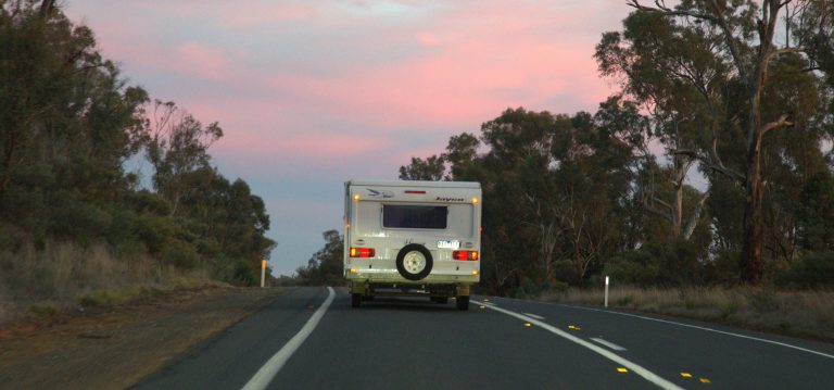 A caravan travelling along a sealed country road at dusk, surrounded by eucalyptus trees under a pink morning sky.